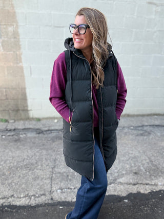Woman wearing a black puffer vest over a purple shirt against a gray wall.